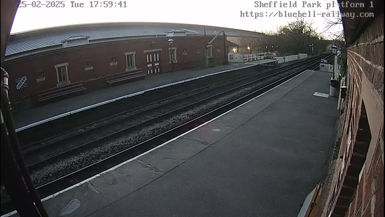 A train platform with two sets of tracks, a brick building, and a dusk sky is visible from a low angle.