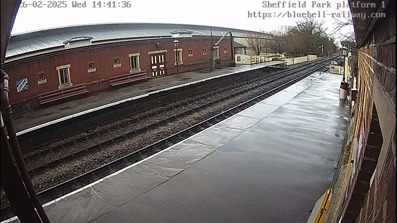 A wet train platform with railroad tracks and a brick building in the background.