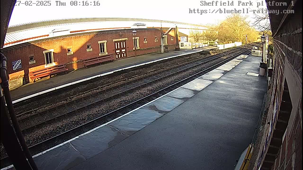 A mostly sunny view of a train platform with a brick building and train tracks.