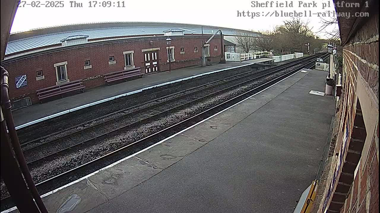 A mostly overcast day reveals a train platform with two sets of tracks leading to a brick building with several windows and benches.