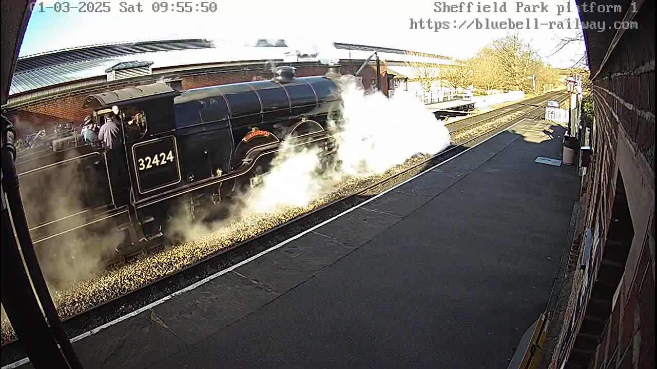 A steam train is departing a railway station on a sunny day, emitting a plume of white smoke.