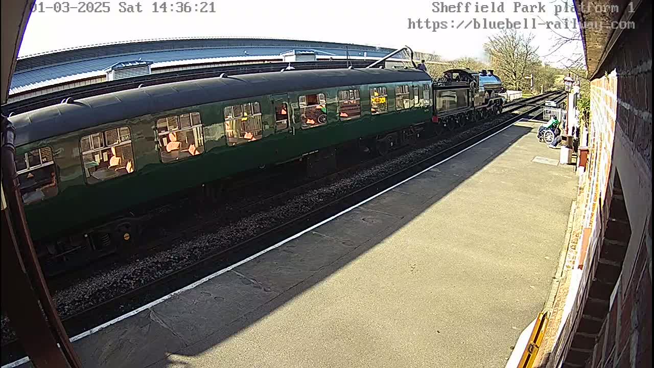 A long, dark-green passenger train is at a station platform on a sunny day.