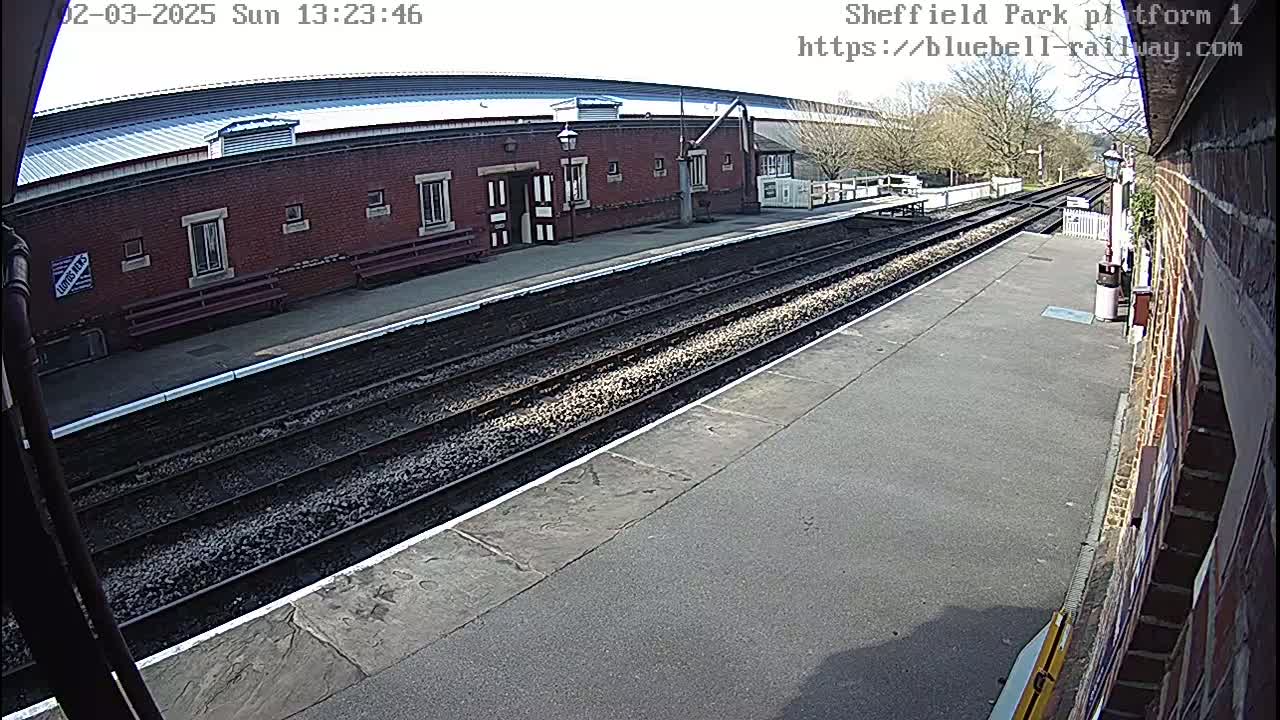 A train platform with a brick building, train tracks, and a sunny day.
