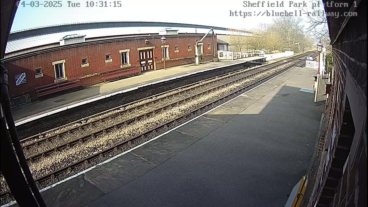 A sunny view of a train platform with a brick building, train tracks, and benches.