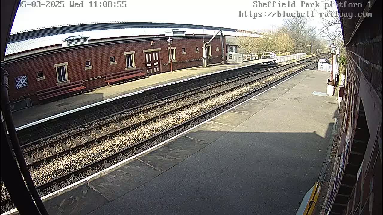 A train platform with multiple train tracks under a sunny sky, features a brick building with benches, and a partially visible adjacent brick wall.