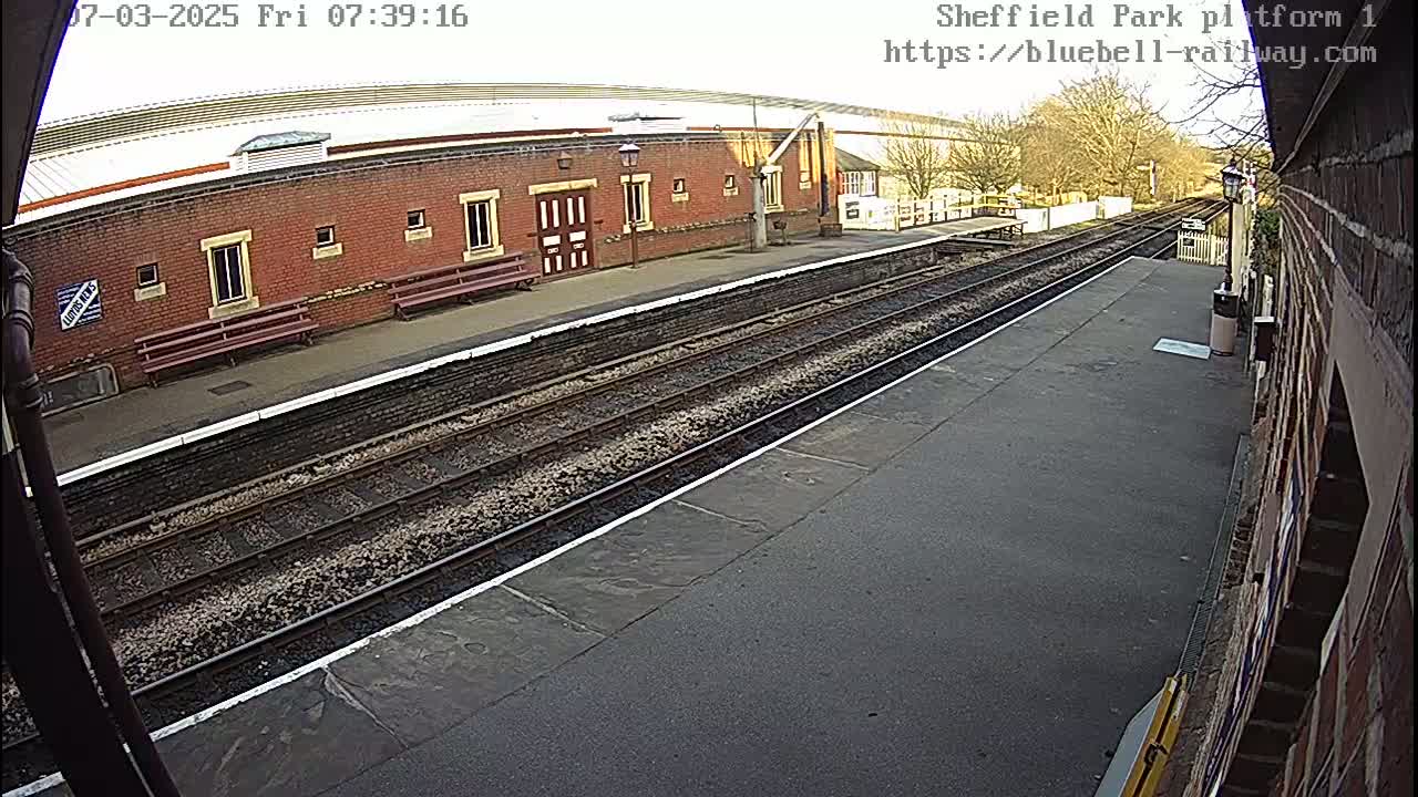 A brick railway station platform with train tracks and benches is visible on a partly sunny day.