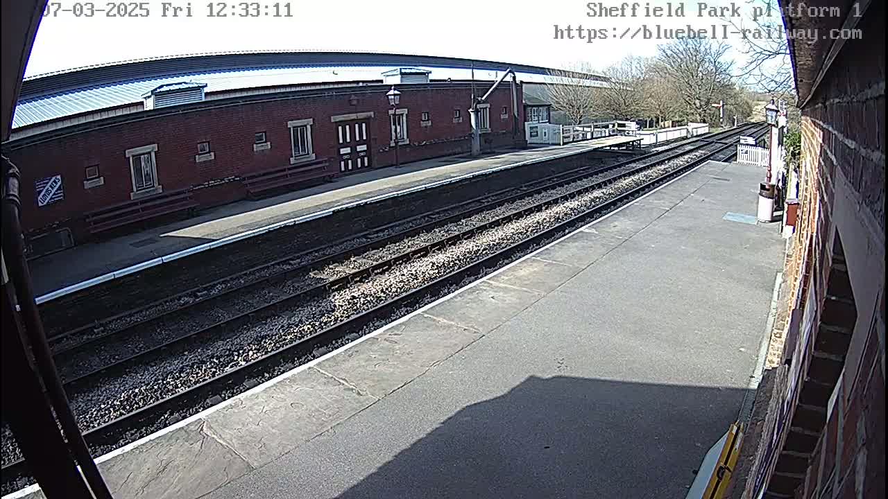 A sunny view of a train station platform with multiple train tracks and a brick building.