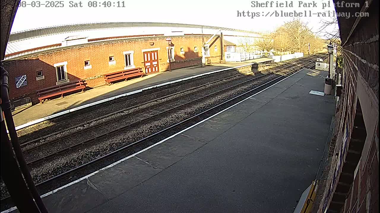A sunny view of a train platform with a brick building, benches, and railway tracks.