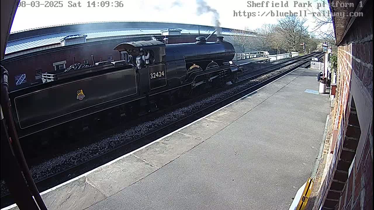 A black steam train departs from a railway station platform on a partly sunny day.