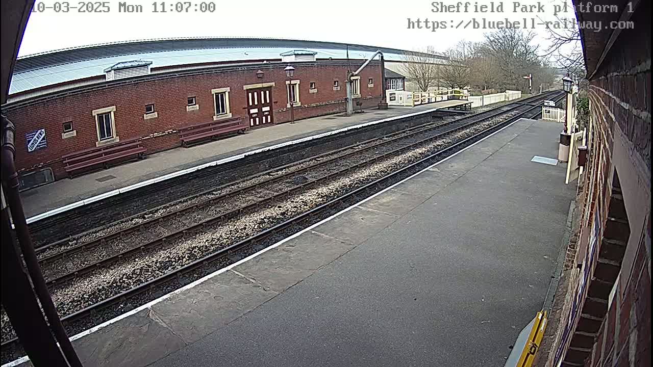 A train platform with a brick building, train tracks, and a mostly clear sky is visible.