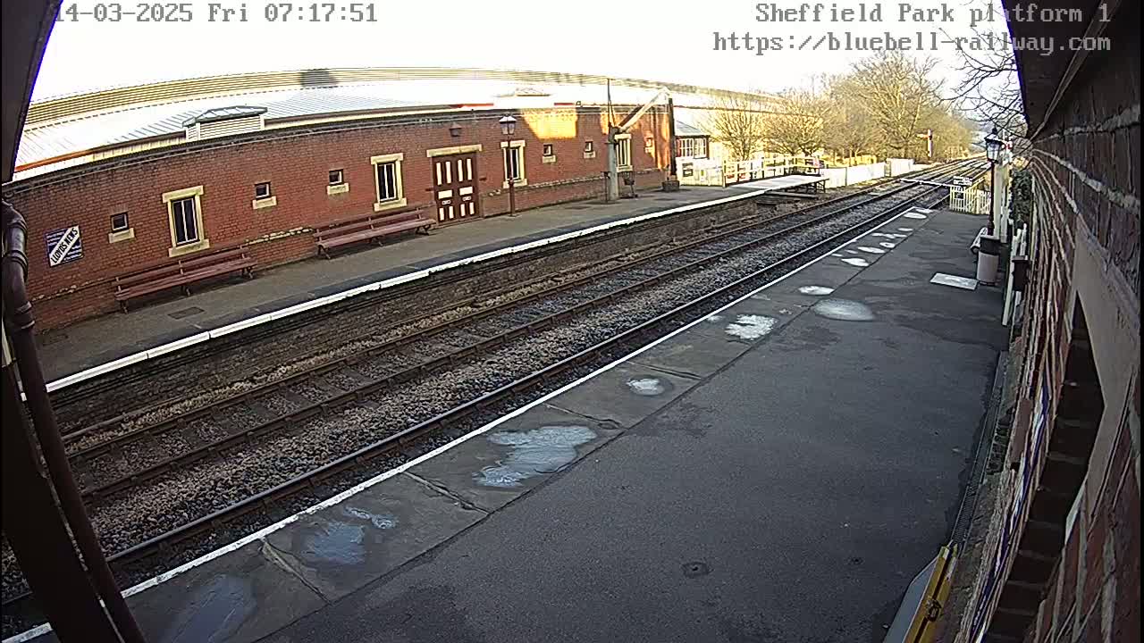 A train platform with a brick building and train tracks under a partly sunny sky.