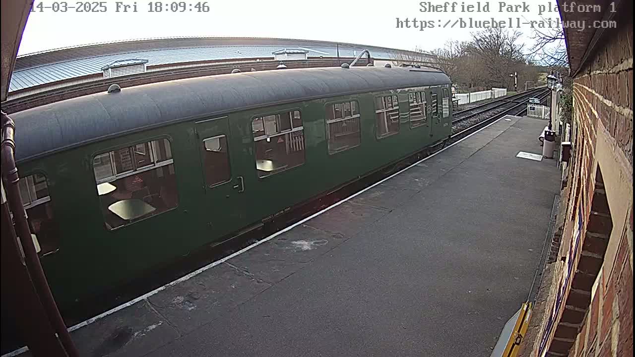 A dark-green passenger train car sits at a railway platform on an overcast day.