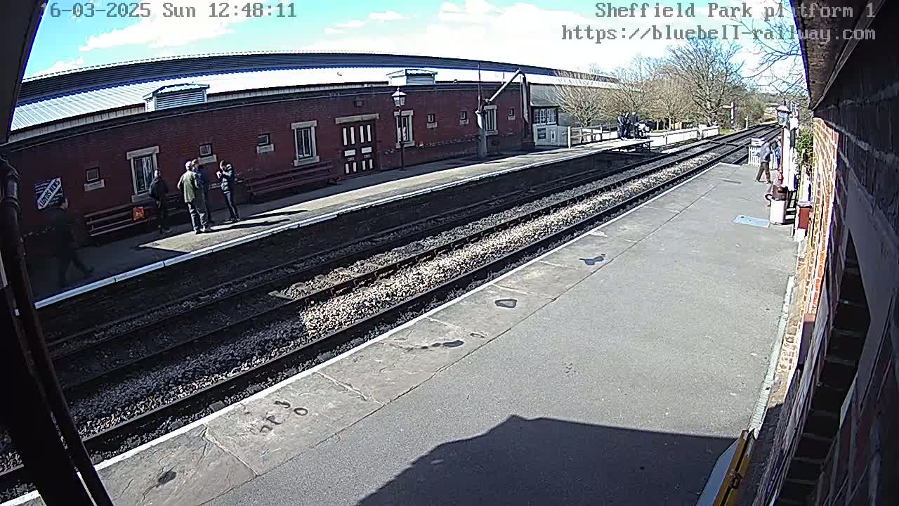 A sunny day at a train station platform with several people standing near a brick building and train tracks.