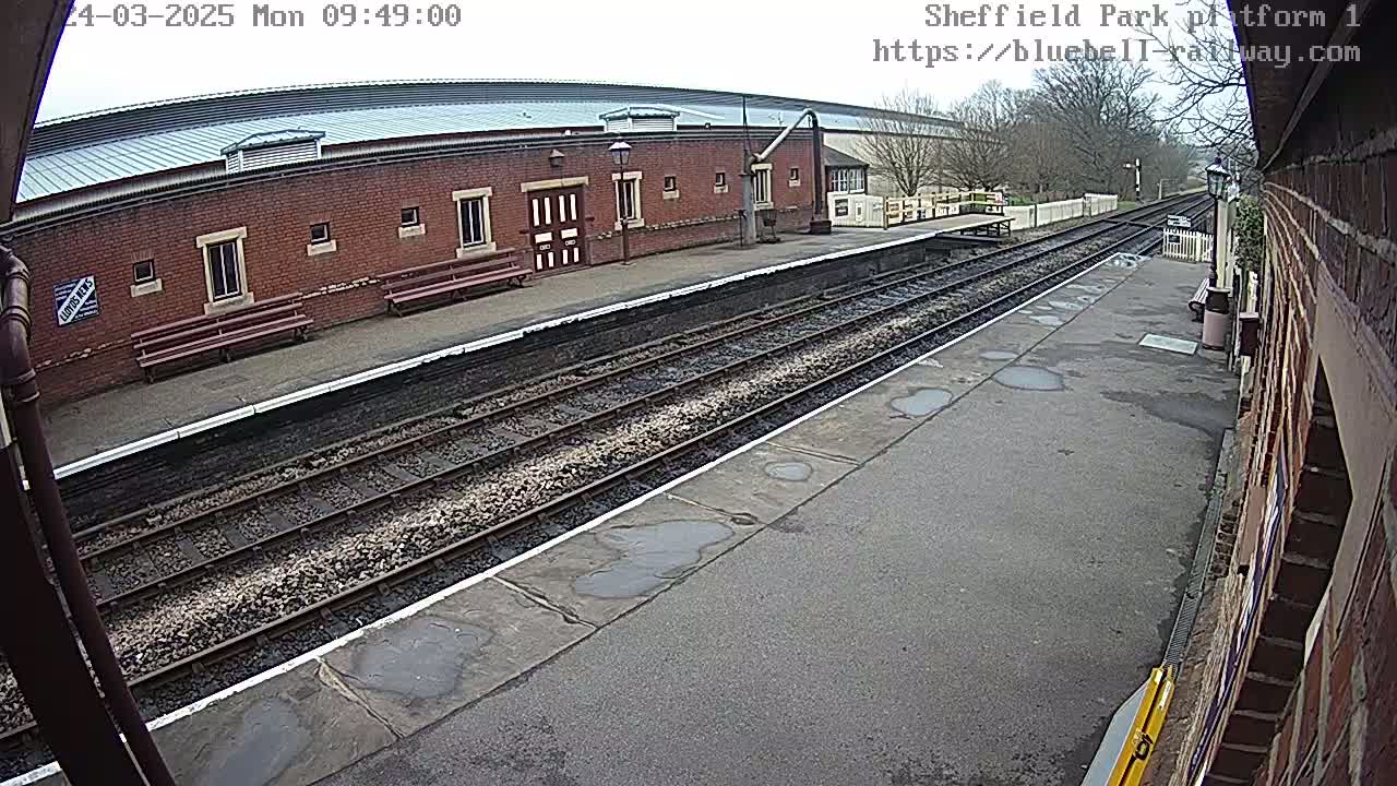 A train platform with a brick building and train tracks, showing some puddles on a cloudy day.