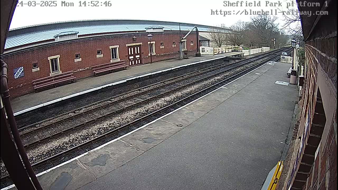 A brick railway station platform with two sets of train tracks under an overcast sky.