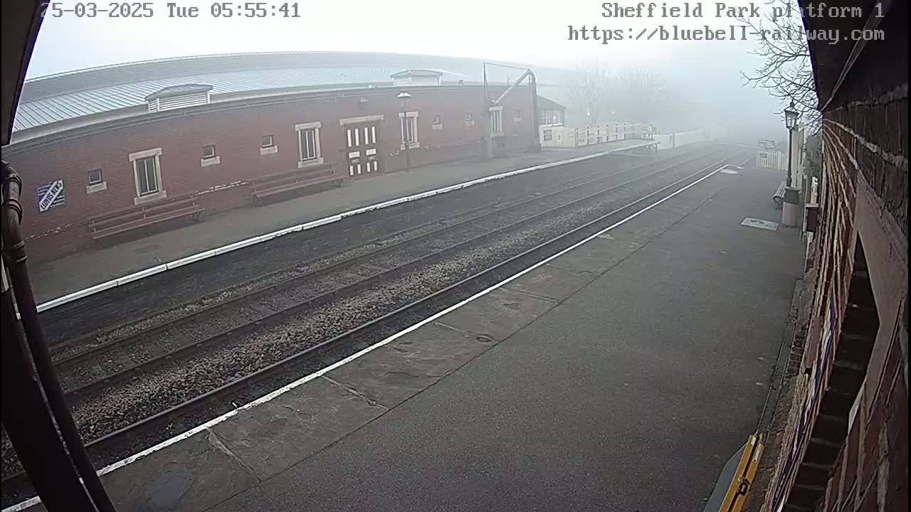 A foggy view of a train platform with two sets of train tracks and a brick building in the background.