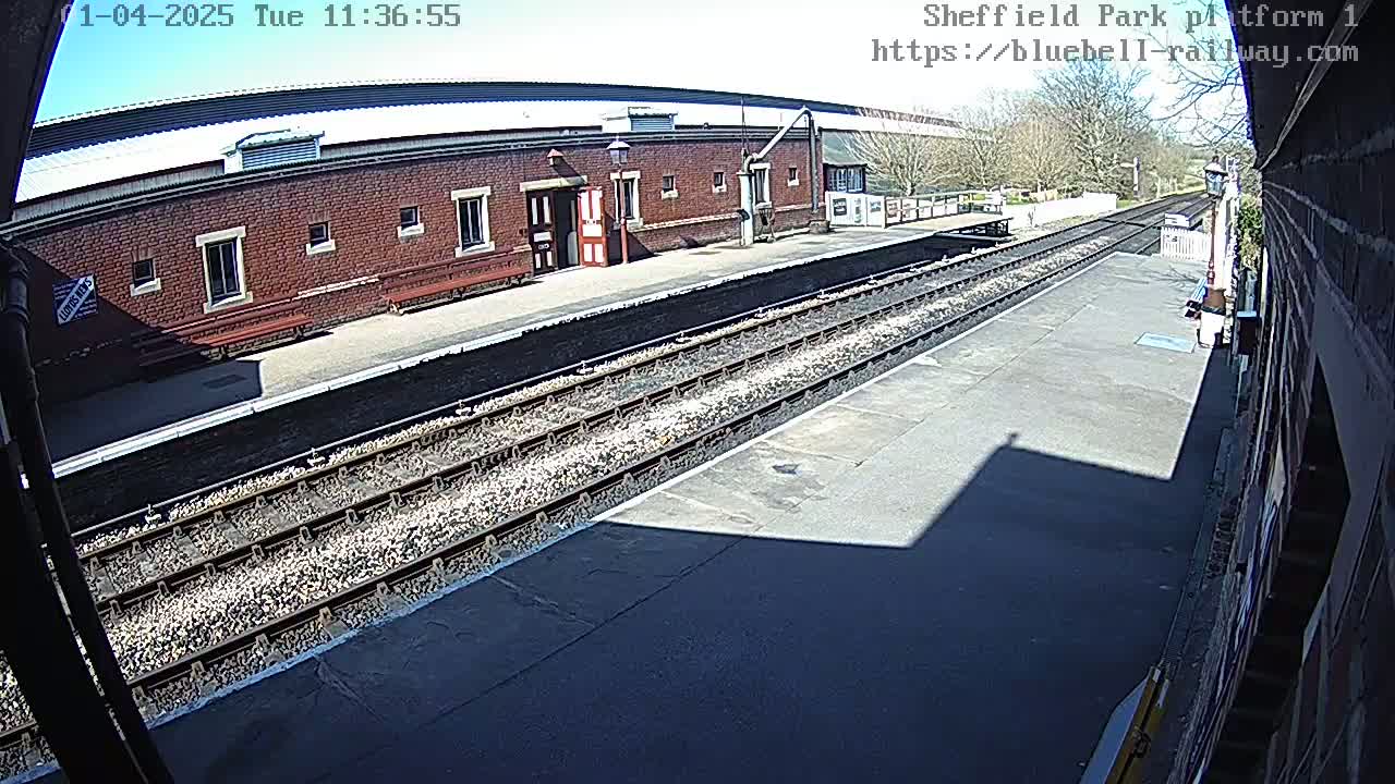 A sunny view of a train platform with a brick building, train tracks, and a long shadow cast across the platform.