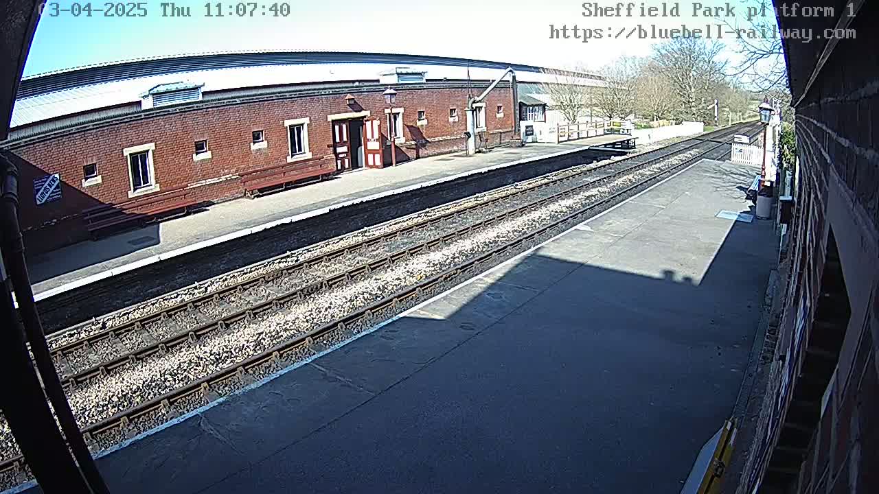 A sunny view of a train platform with a brick station building and multiple train tracks.