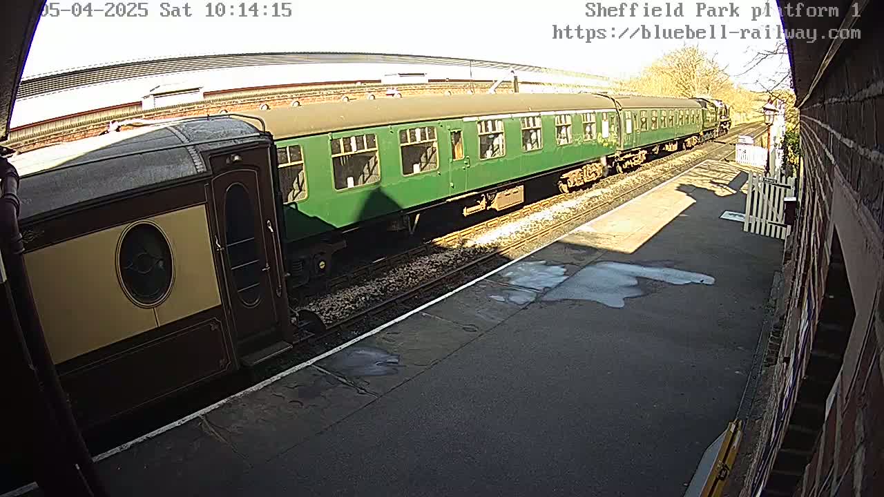 A long green passenger train and a shorter cream and brown passenger car are on a train track on a sunny day.