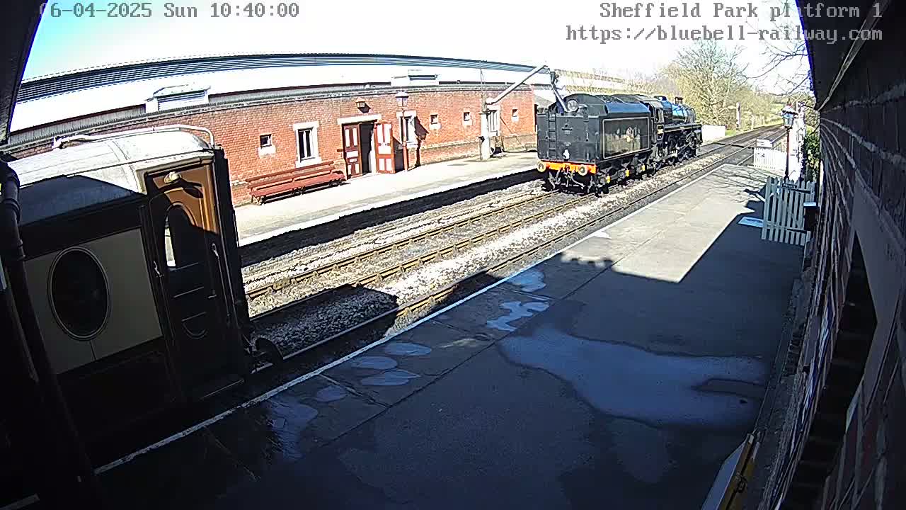 A black steam train and a cream and brown passenger car are at a railway station platform on a sunny day.