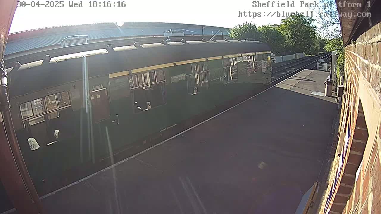 A dark green passenger train car is parked at a train station platform on a sunny day.