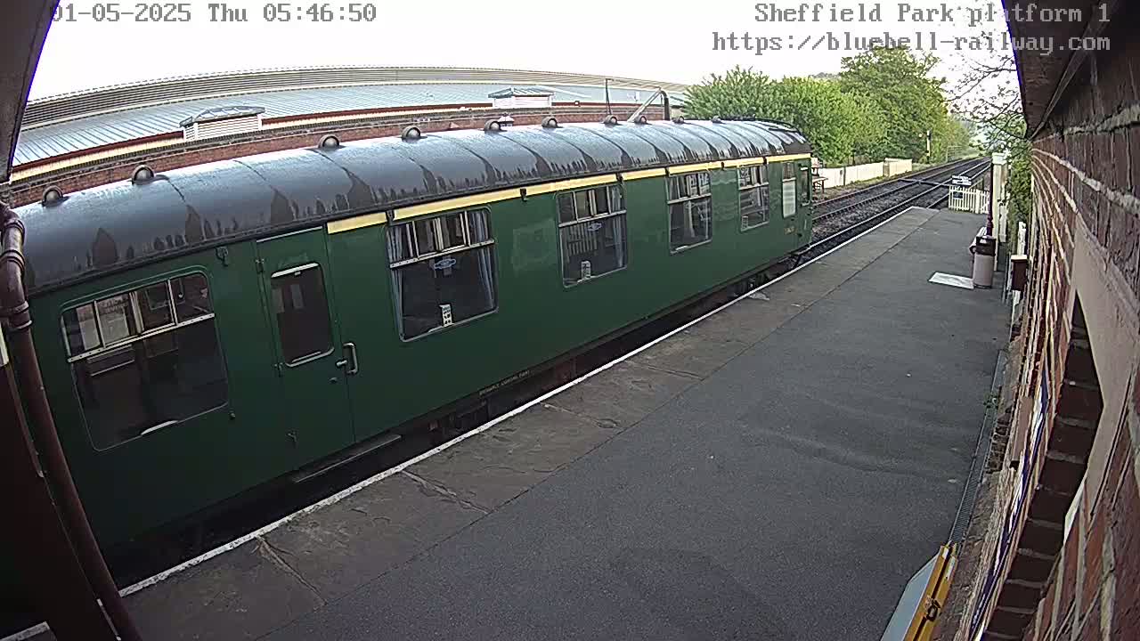 A dark-green passenger train car sits at a train station platform on an overcast day.