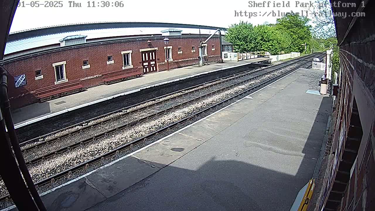 A train platform with a brick building and train tracks under a mostly sunny sky.