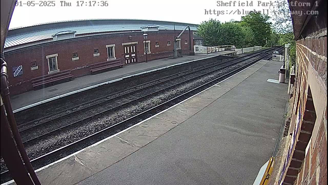 A mostly sunny view of a train platform with two train tracks, a brick building, and benches.