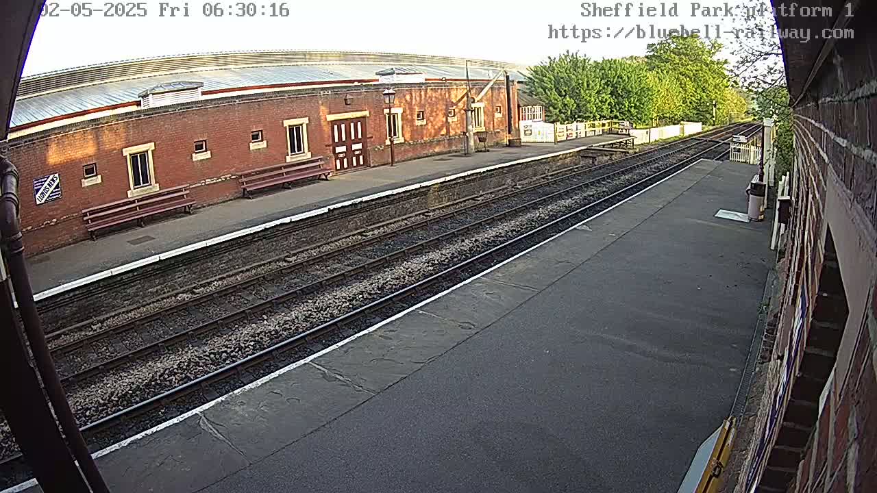 A brick train station building with benches sits alongside train tracks on a partly sunny morning.