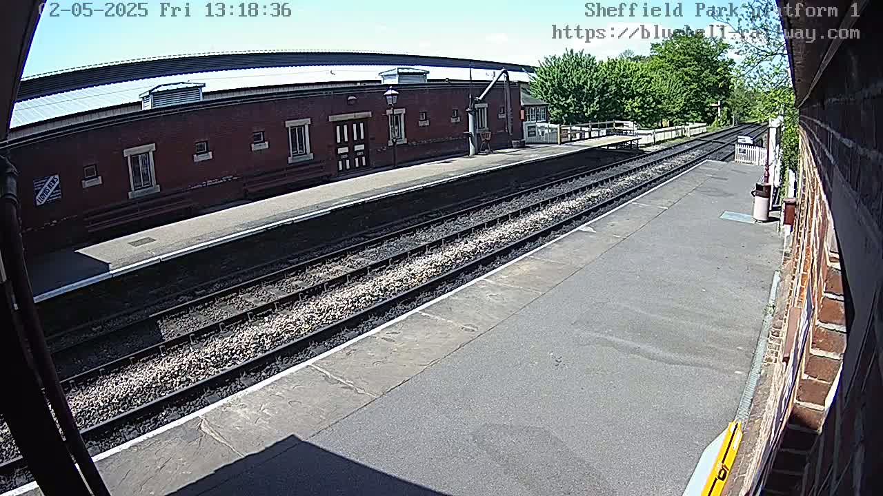 A sunny view of a train platform with two parallel train tracks, a brick building, and some trees in the background.