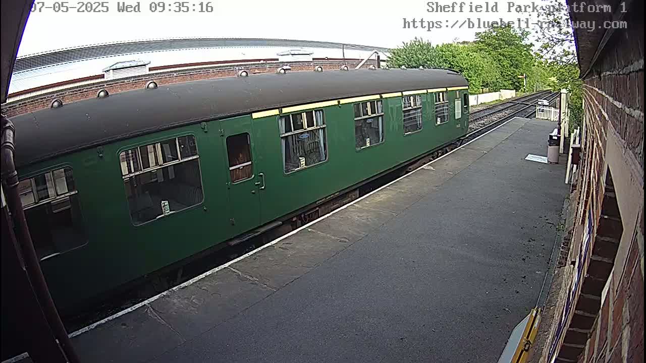 A dark-green passenger train car sits at a station platform on an overcast day.