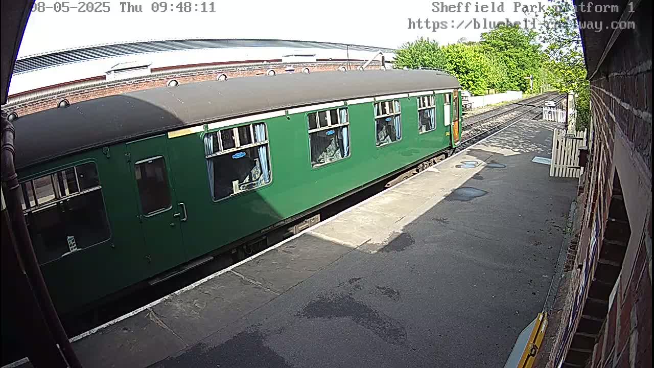 A dark green passenger train is parked at a sunny train station platform.