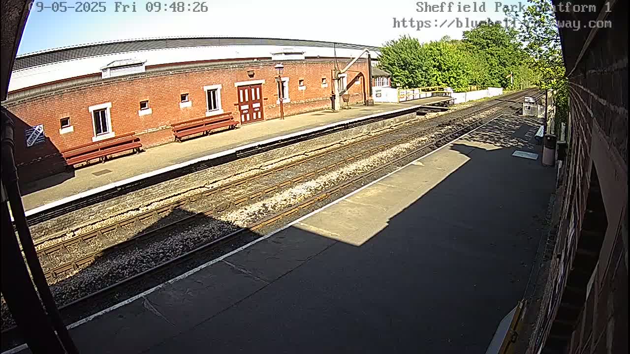 A sunny view of a train platform with a brick building, benches, and train tracks.