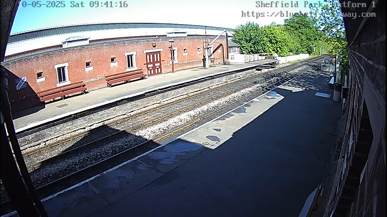 A sunny view of a train platform with a brick building, benches, and train tracks.