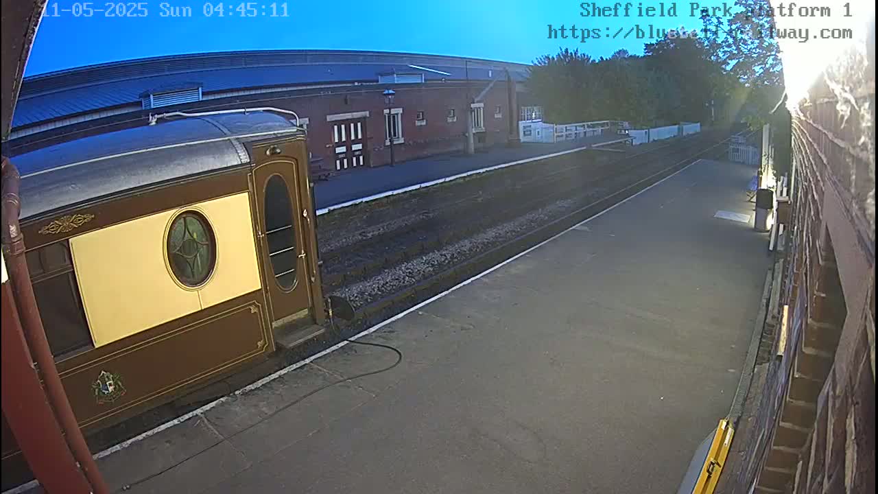 A brown and tan passenger rail car sits at a train station platform under a clear, early morning sky.