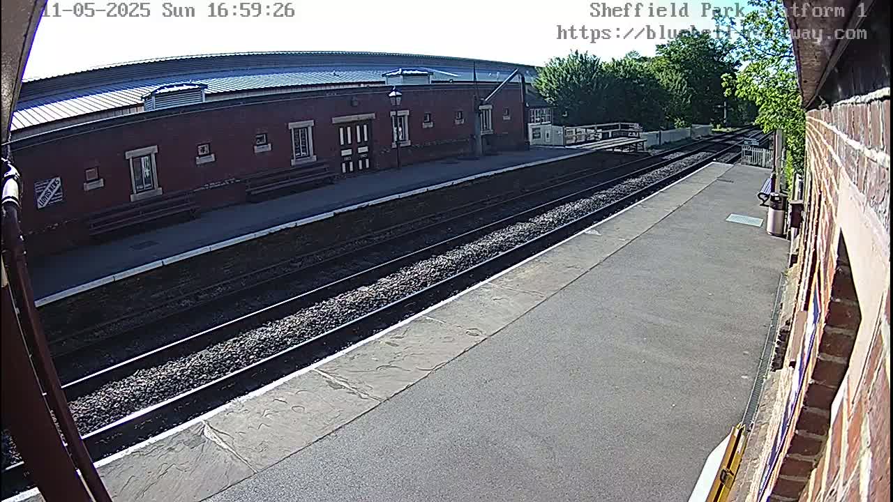 A sunny day at a train station shows a brick building, train tracks, and a platform.
