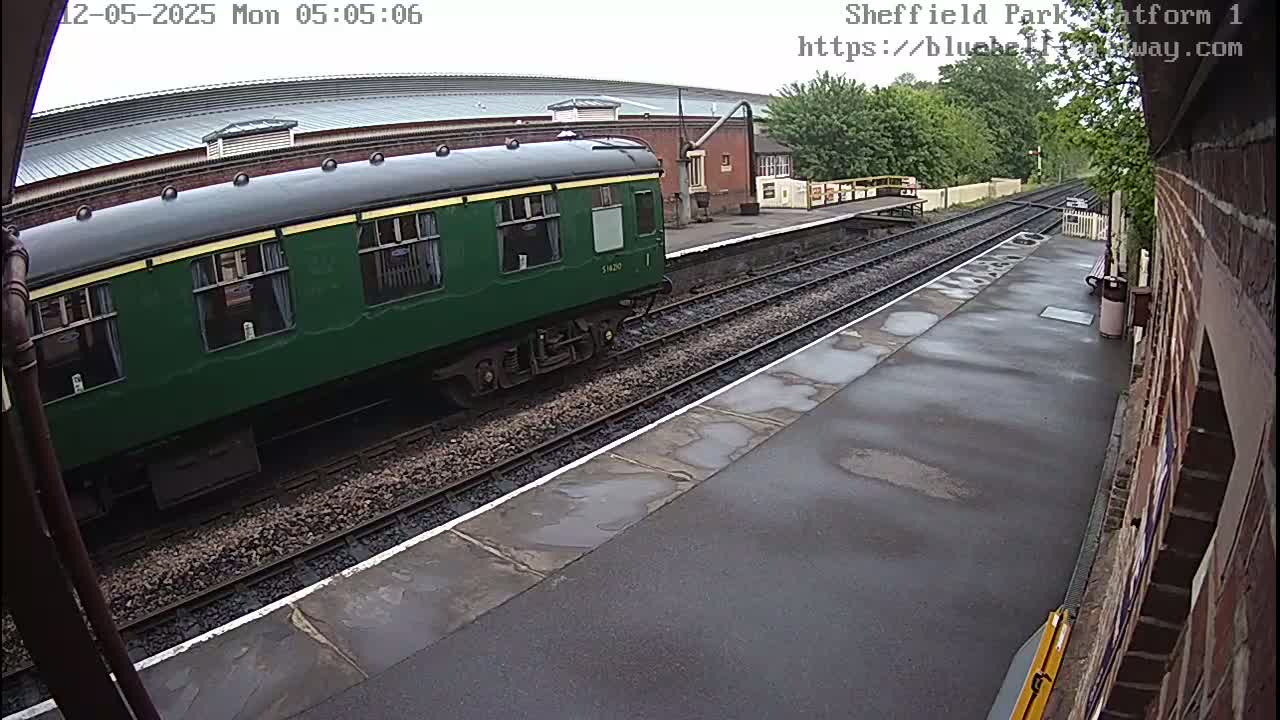 A dark-green railway carriage is parked at a train station platform on a wet, overcast day.