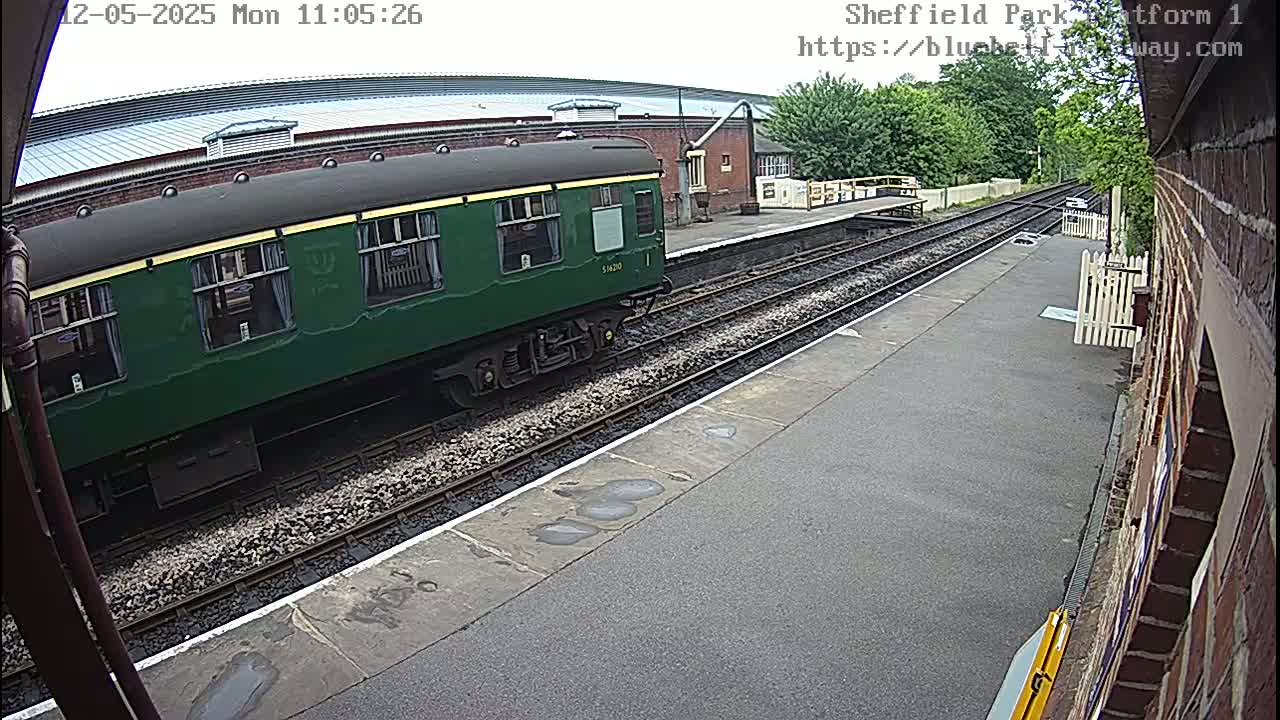 A dark-green passenger train car sits on a train track next to a platform on a partly sunny day.
