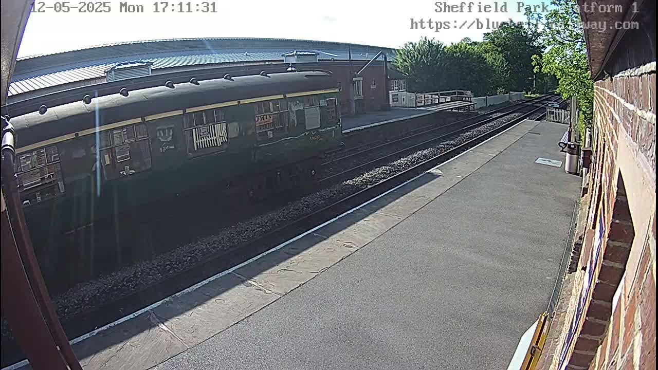 A dark-green passenger train is at a station platform on a sunny day.