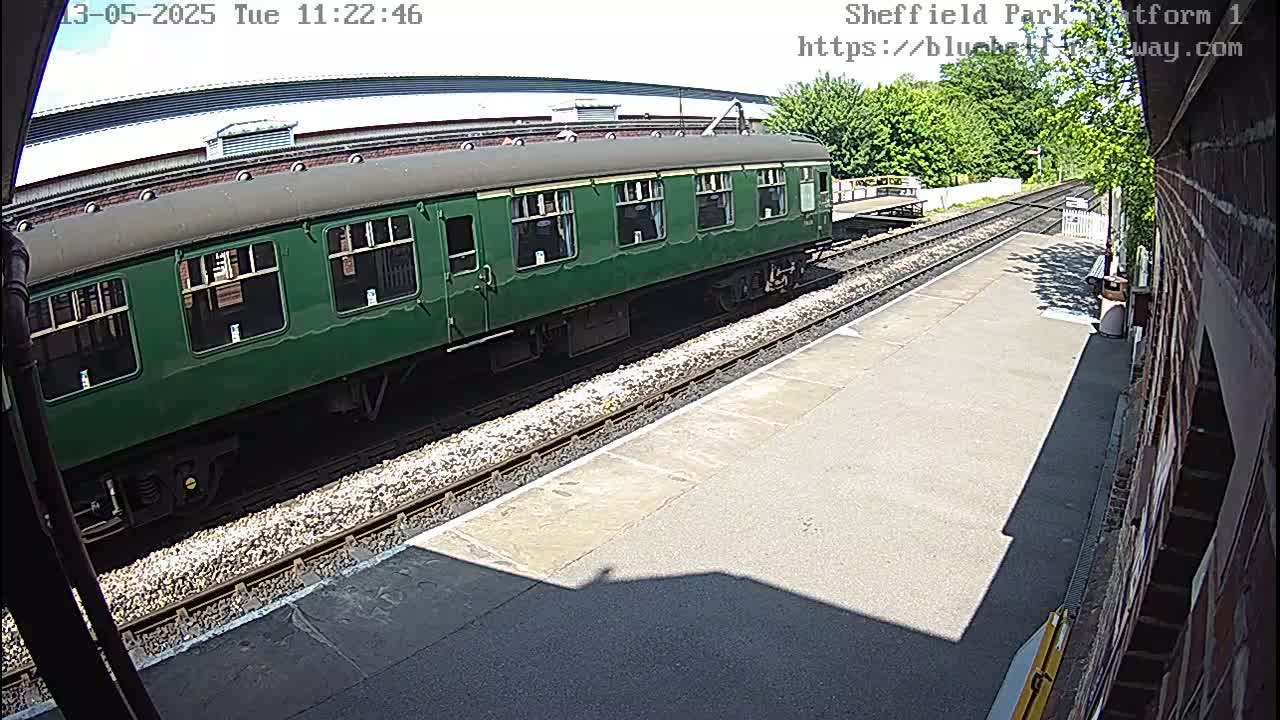 A long, dark-green train car sits on a sunny train platform next to train tracks.