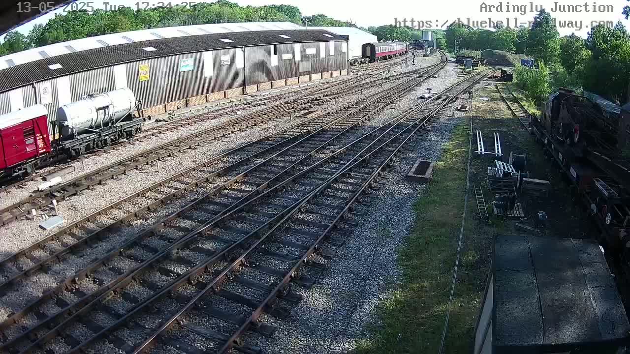 A sunny day at a railway yard shows multiple train cars and locomotives on several tracks next to a long building.