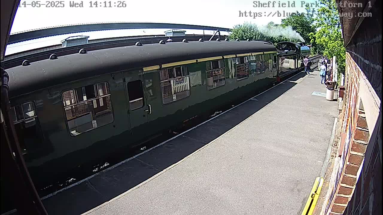 A dark green passenger train car sits at a station platform on a sunny day, with several people standing nearby.