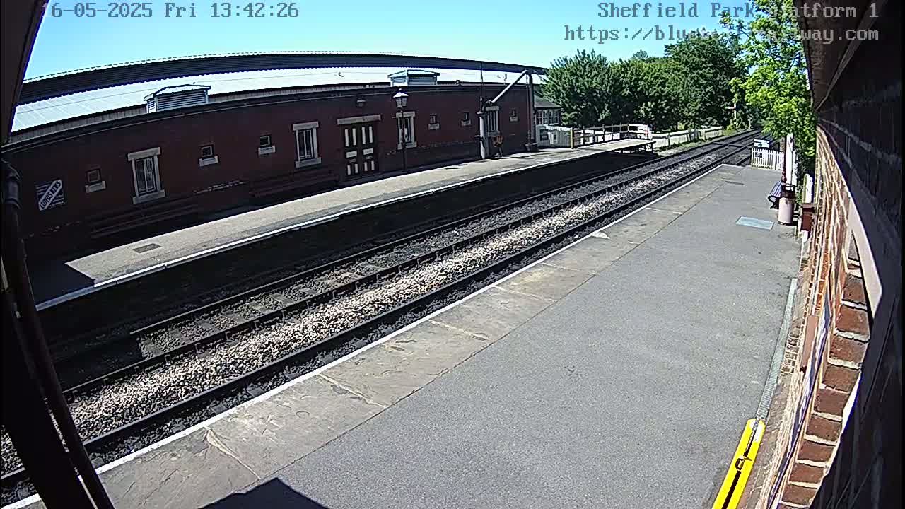 A sunny view of a train platform with two sets of train tracks and a brick building in the background.