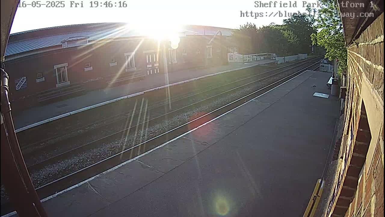 A sunny view of a train station platform with train tracks and a brick building in the background.