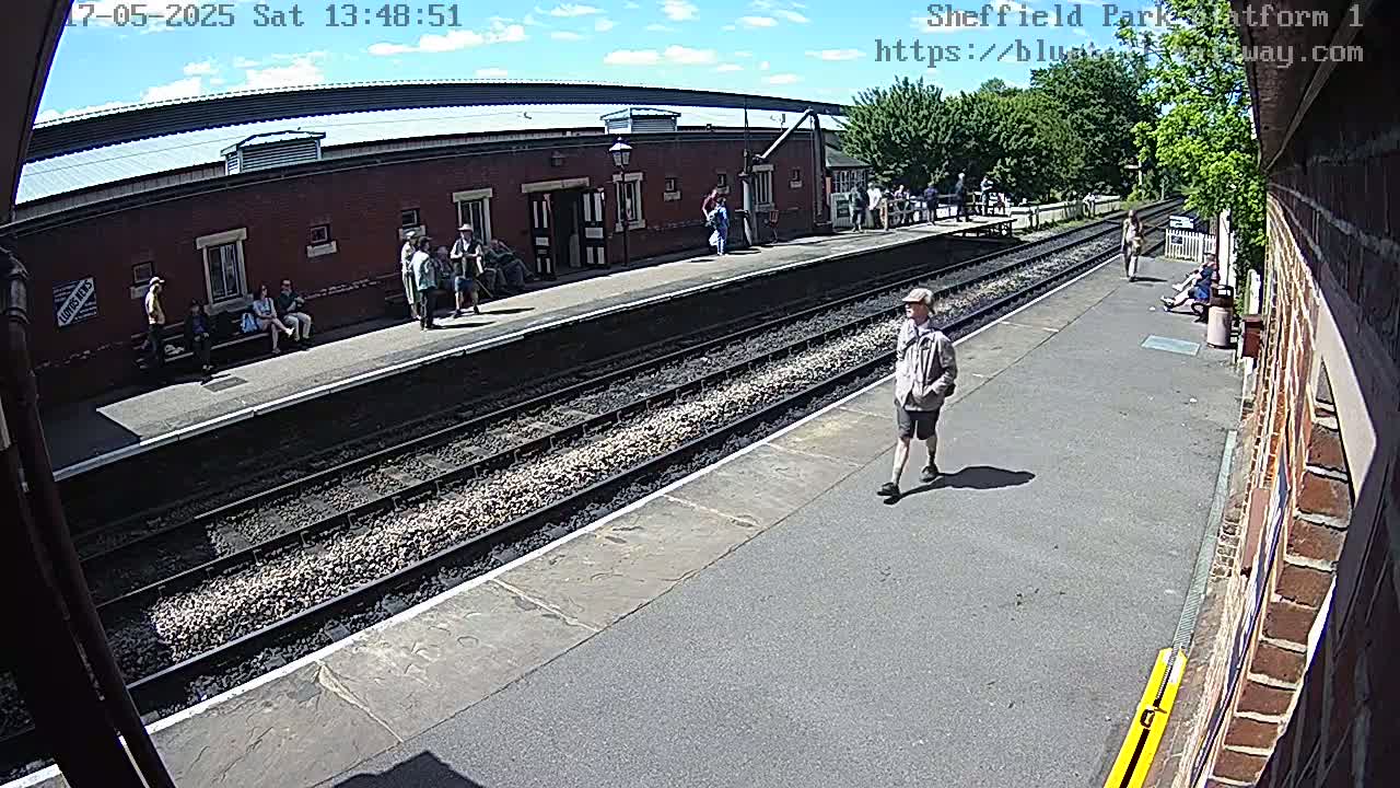 A sunny day at a train station platform shows several people waiting near train tracks and a brick building.