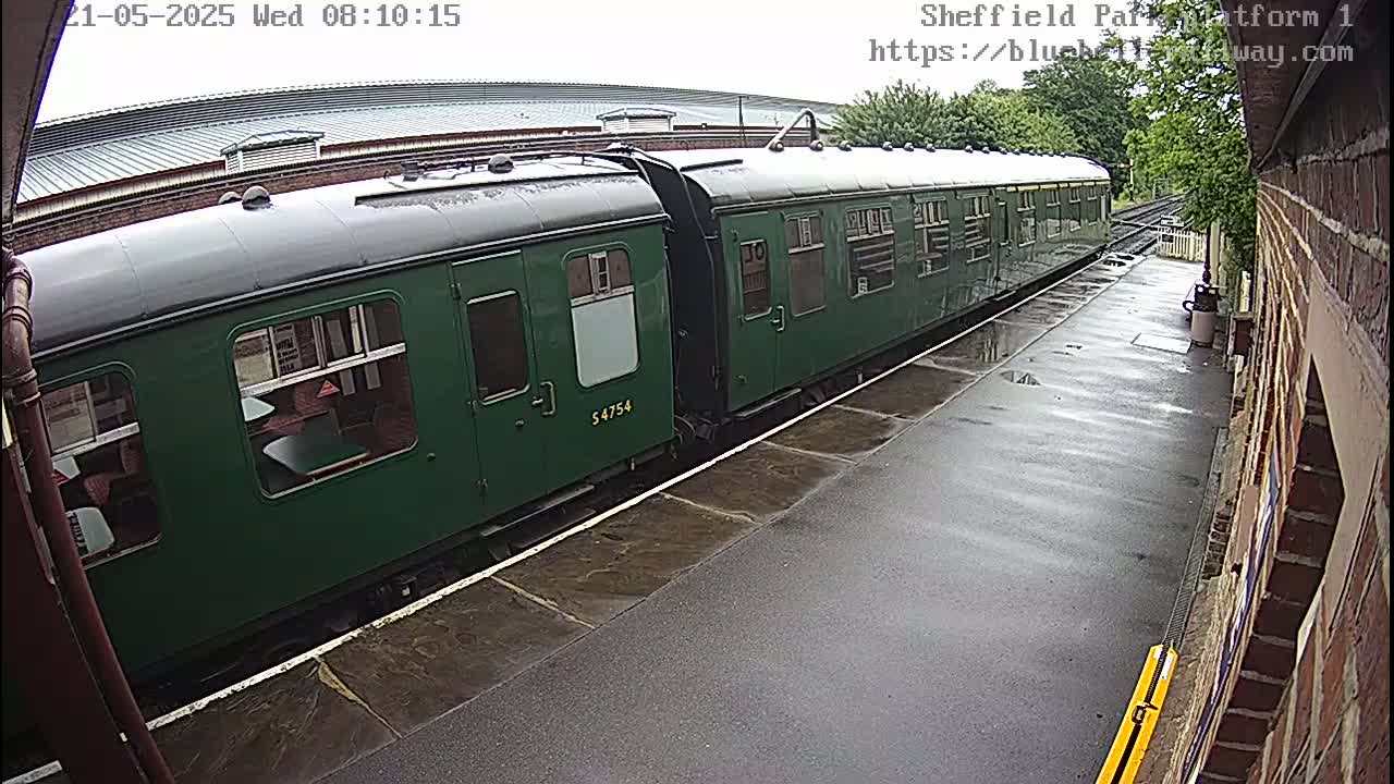 A long, dark-green passenger train sits on a wet train platform next to a brick building.