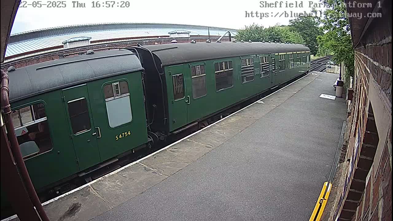 A long, dark-green passenger train is at a station platform on an overcast day.