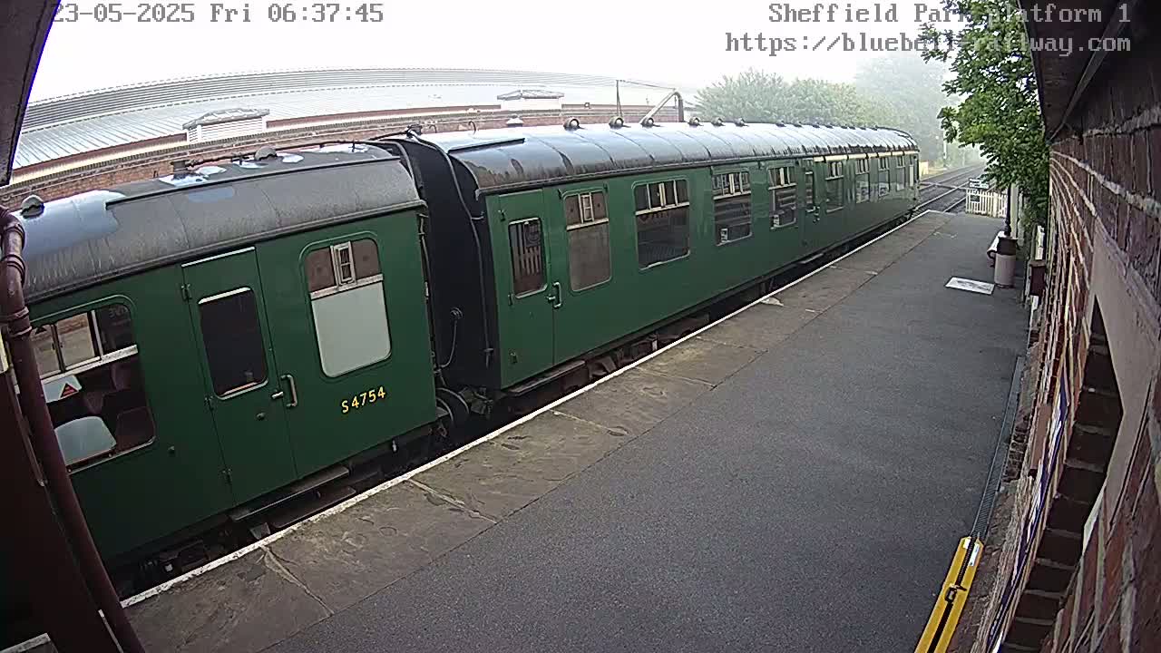 A long, dark-green passenger train is at a station platform on a misty morning.