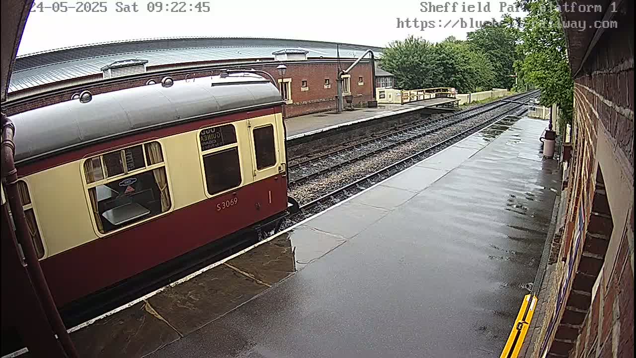 A maroon and cream passenger train car sits at a rain-slicked railway platform next to a brick building.