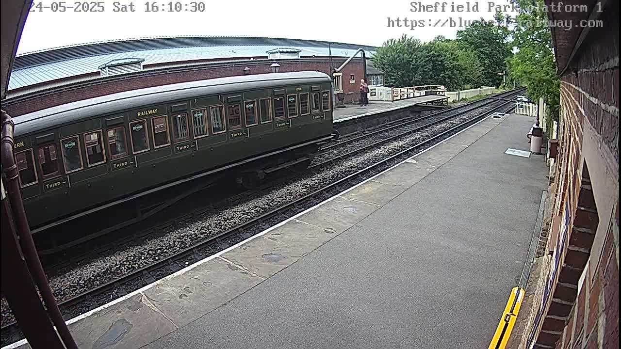 A dark-green passenger train car is partially visible at a train station on a partly cloudy day.